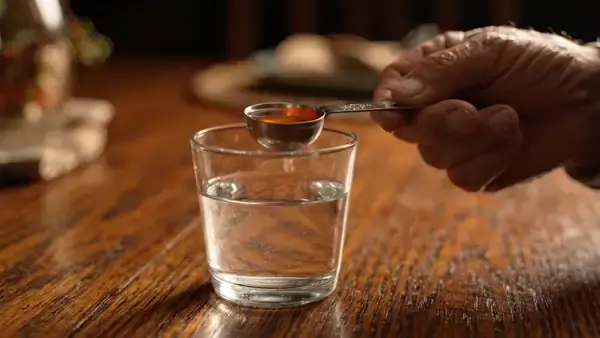 A measuring teaspoon of pale orange liquid being tipped into a glass of water on a supper table