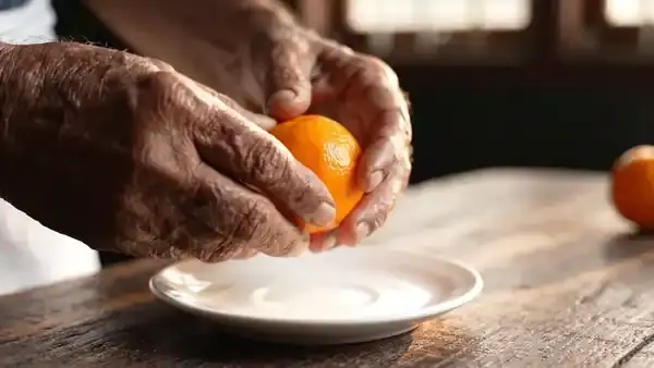 An older man's hands peeling a tangerine over a white saucer on a kitchen table